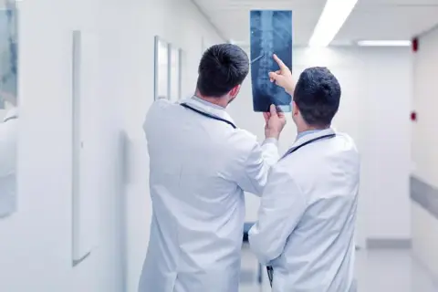Dwóch lekarzy od kręgosłupa analizuje zdjęcie rentgenowskie kręgosłupa w szpitalnym korytarzu.