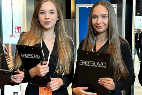 Dwie uśmiechnięte hostessa z agencji PROPROMO trzymają teczki z logo firmy.