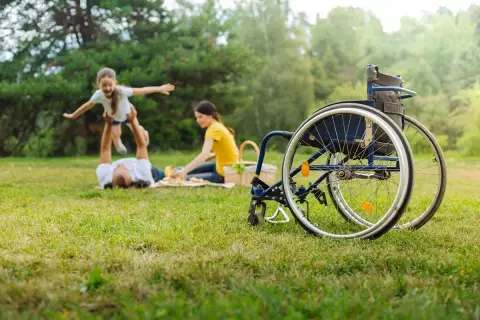 Rodzina na pikniku, ojciec podrzuca córkę, matka przygotowuje jedzenie. Obok wózek inwalidzki, symbol wsparcia i świadczenie pielęgnacyjne 2026.