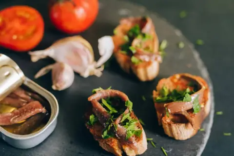 Otwarta puszka anchois, bruschetty z anchois i natką pietruszki, pomidory i czosnek na ciemnym tle.