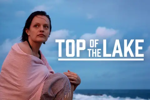 Kobieta owinięta w różowy ręcznik siedzi na plaży, patrząc w dal. Na niebie widnieje napis "TOP OF THE LAKE".