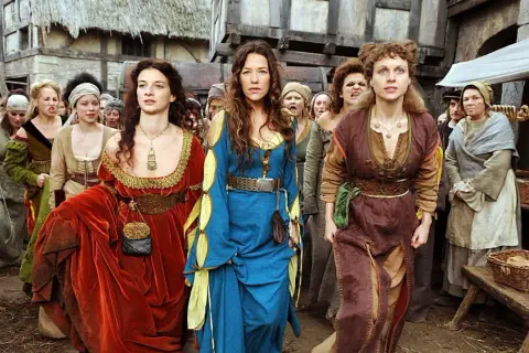 Drei Frauen in historischen Gewändern, die an eine "die wanderhure reihenfolge" erinnern, gehen durch eine mittelalterliche Gasse.