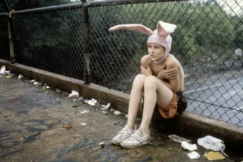 Bambino con cappello da coniglio seduto su un marciapiede sporco, un'immagine che evoca la trama di Gummo.