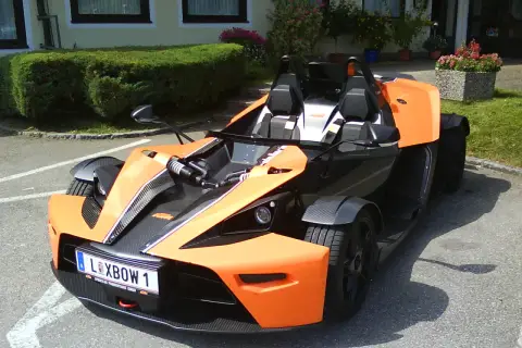 Pomarańczowy KTM X-Bow z czarnymi akcentami i tablicą rejestracyjną "XBOW 1" stoi na parkingu przed budynkiem z zielonym żywopłotem.