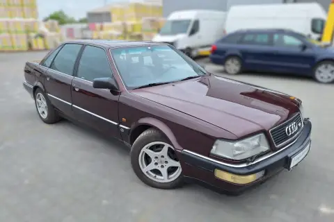 Bordowy Audi V8 z lat 90. na parkingu, w tle białe vany i niebieski kombi.