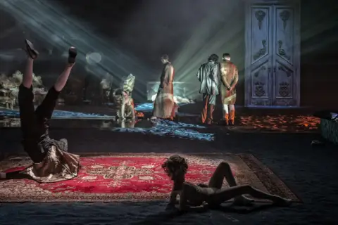 Scenograf tworzy surrealistyczny świat. Na scenie akrobata, dwie postacie w strojach z epoki i wielkie drzwi.