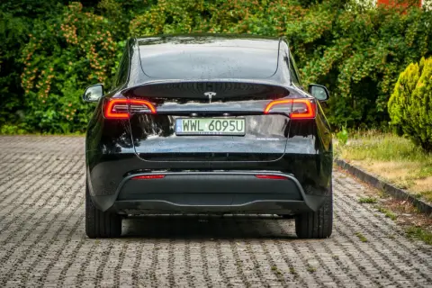 Czarna Tesla z napędem dual motor, z tablicą rejestracyjną WWL 6095U, zaparkowana na kostce brukowej wśród zieleni.