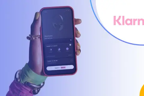 Dłoń trzyma smartfon z aplikacją Klarna, umożliwiającą zakup słuchawek na raty.