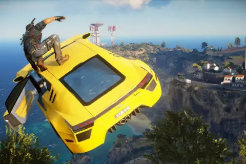 Rico z Just Cause 5 skacze z żółtego supersamochodu nad klifem, w tle widać śródziemnomorską wioskę i morze.