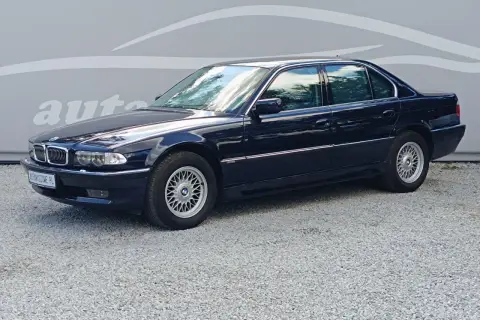 Ciemnoniebieskie BMW 7 E38 z lat 90. stoi na żwirowym podjeździe, z odbiciem chmur w szybach.