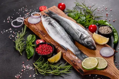 Dwie świeże ryby bass na desce, otoczone cytryną, limonką, pomidorkami, cebulą, rozmarynem i pieprzem.