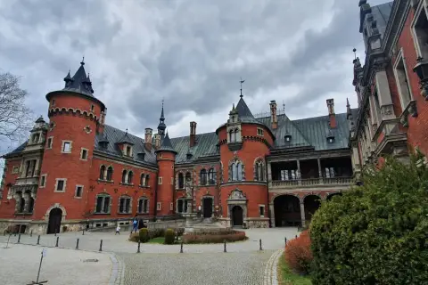 Pławniowice atrakcje: okazały zamek z czerwonej cegły z wieżami i arkadowym gankiem, otoczony brukowanym dziedzińcem i zielenią.