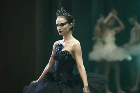 Natalie Portman jako Czarny Łabędź w koronie i czarnej tutu, z innymi tancerkami w tle.