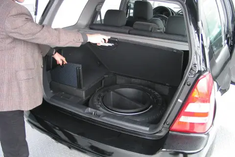 Mężczyzna otwiera bagażnik, pokazując schowek na koło zapasowe i subwoofer. Awaryjne otwieranie bagażnika może być konieczne w razie problemów.