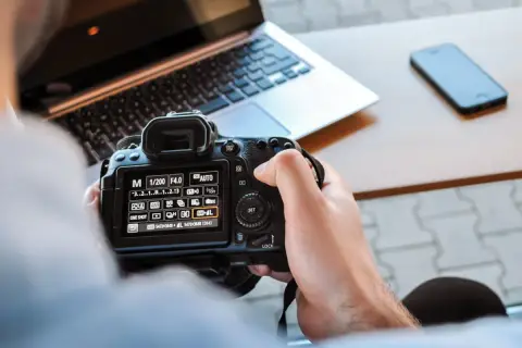 Ręce trzymają aparat, na ekranie ustawienia manualne. Nauka fotografii w praktyce, obok laptop i telefon.