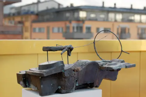 En abstrakt skulptur i mörk metall, placerad på ett vitt podium. I bakgrunden syns byggnader och en gul vägg, kanske vid museum Vasaparken.