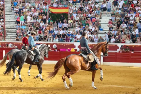 Dos jinetes, uno con chaquetilla roja y otro con azul, montan caballos en una plaza de toros. Bernardino Piriz, con sombrero, observa la escena.