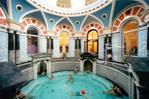 Wnętrze z basenem, gdzie ludzie relaksują się w gorącym źródle. Architektura jest bogato zdobiona, z niebieskimi i pomarańczowymi detalami.