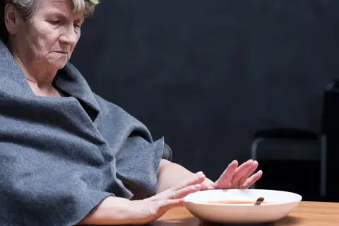 Starsza kobieta w szarym szalu odwraca się od talerza z zupą, wyraźnie odczuwając brak apetytu.