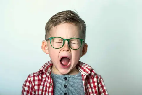 Niño con gafas verdes y camisa a cuadros, con los ojos cerrados y la boca abierta, expresando una emoción intensa, como podría ocurrir en el síndrome de Tourette en niños.