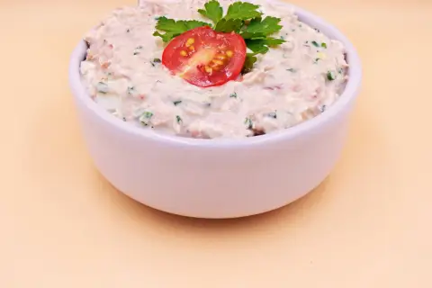 Pyszna pasta z tuńczyka i jajka, udekorowana pomidorkiem i natką pietruszki, w białej miseczce.