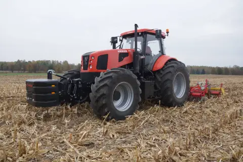 Czerwony Ursus 28014 z obciążnikiem z przodu i pługiem z tyłu pracuje na polu po żniwach.