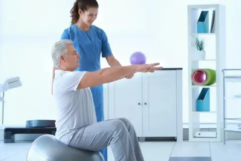 Fizjoterapeuta pomaga starszemu mężczyźnie w ćwiczeniach izometrycznych na piłce gimnastycznej.