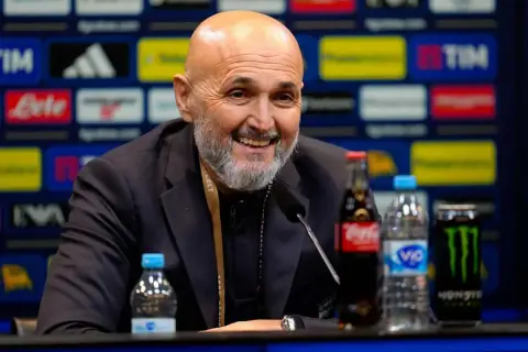 Uśmiechnięty trener Juventusu, z siwą brodą, siedzi przy stole z napojami.