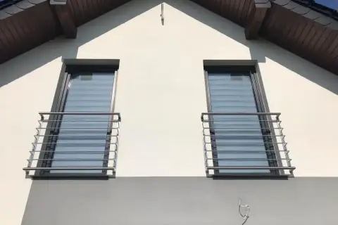 Nowoczesny montaż balkonu francuskiego na białej elewacji domu. Dwa okna z balustradami ze stali nierdzewnej.