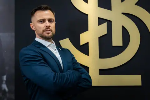 Mężczyzna w garniturze, trener ŁKS Łódź, stoi przed złotym logo klubu.