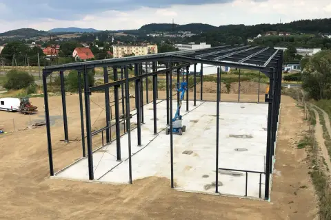 Stalowa konstrukcja hali przemysłowej, gotowa na montaż płyt warstwowych. W tle krajobraz z pagórkami i zabudowaniami.