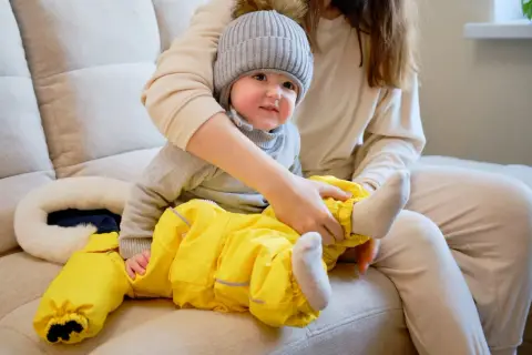 Mama ubiera dziecko w żółty kombinezon i czapkę. Wskazówki, jak ubrać dziecko odpowiednio do temperatury, pomogą Ci przygotować malucha na spacer.