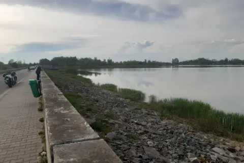 Nad bralinem, gdzie woda odbija chmury, stoi człowiek. Obok motocykle i ścieżka z kostki brukowej.