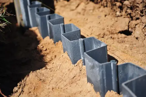 Szare, metalowe elementy, przypominające małe **larseny**, są wkopane w piasek, tworząc linię.