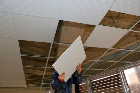 Pracownik montuje płyty do ocieplenia sufitu podwieszanego. Widać odsłonięte elementy konstrukcji i stare betonowe strop.