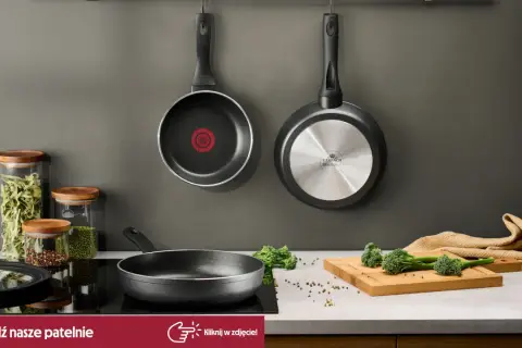 Dwie patelnie wiszą na ścianie, jedna stoi na płycie indukcyjnej. Obok słoiki z makaronem i przyprawami, deska z brokułami. To najlepsza patelnia do Twojej kuchni!