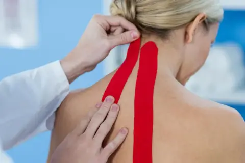 Fizjoterapeuta aplikuje czerwony taping medyczny na kark pacjentki, wspierając mięśnie i poprawiając krążenie.