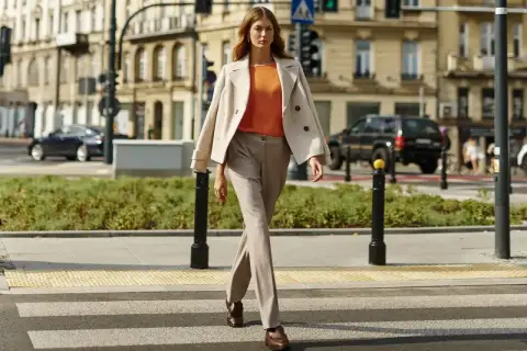 Elegancka kobieta w beżowym płaszczu i pomarańczowej bluzce przechodzi przez pasy. Stylowe stylizacje do beżowego płaszcza na co dzień.