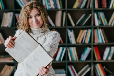 Kobieta z książką w bibliotece. Dowiedz się, jakie są zasady dystrybucji książek do bibliotek publicznych i szkolnych.