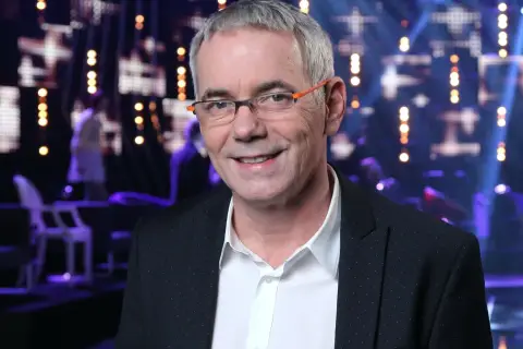 Uśmiechnięty Robert Janowski w okularach na tle sceny. Wiele osób zastanawia się, ile lat ma Robert Janowski, ale jego wiek nie jest widoczny na zdjęciu.