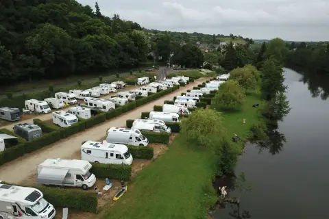 Vue aérienne d'une aire de camping-car gratuite en bord de mer normandie, avec de nombreux véhicules stationnés près d'une rivière bordée d'arbres.
