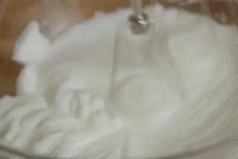 Un batteur électrique fouette un œuf blanc en neige dans un bol en verre, créant une texture aérienne et mousseuse.