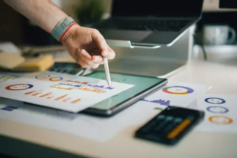 Ręka z rysikiem analizuje wykresy na tablecie. W tle laptop i dokumenty. Broker ubezpieczeniowy pracuje nad analizą danych.
