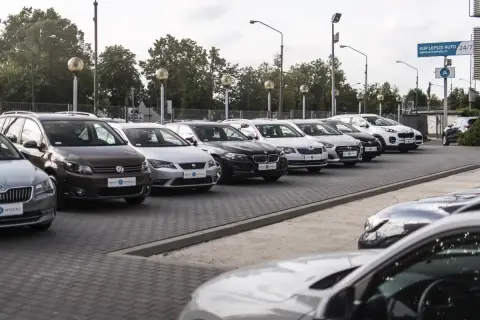 Rząd samochodów na placu, w tym brązowy Volkswagen i biały Kia Sportage. W tle drzewa i słupy oświetleniowe. To idealne miejsce, by znaleźć swój wymarzony kinto one.