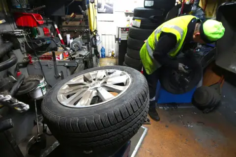 Mechanik w kamizelce odblaskowej pracuje przy kole. Słychać buczenie opon podczas jazdy, gdy pracownik wymienia oponę.
