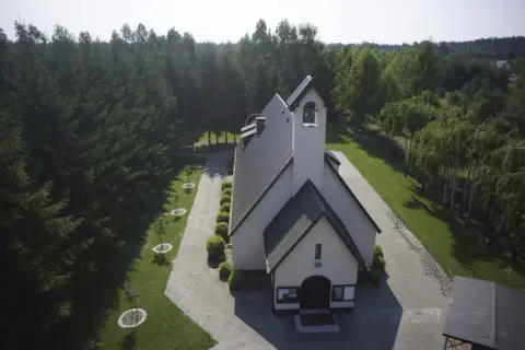 Kościół otoczony lasem, z wieżą i dzwonami. Wokół zadbane alejki i młode drzewka, tworzące urokliwe przypki.