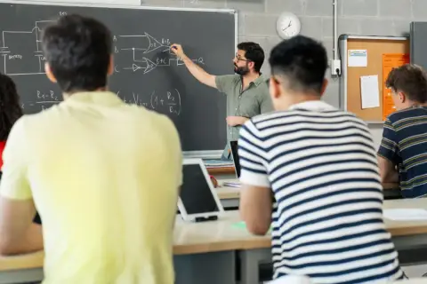 Nauczyciel tłumaczy schemat na tablicy, studenci słuchają. To część jego pensum, by przekazać wiedzę.