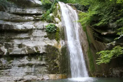 Cascata fragorosa su rocce stratificate, circondata da vegetazione lussureggiante. L'acqua cheta scende impetuosa, creando un velo bianco.