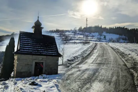 Kamienna kapliczka na zboczu ośnieżonej koskowa góra, z drogą wijącą się w słońcu.
