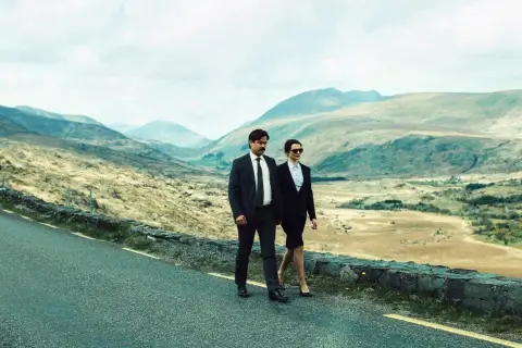 Coppia in abito cammina su strada panoramica, paesaggio montuoso. Un'immagine che evoca la recensione di "The Lobster".